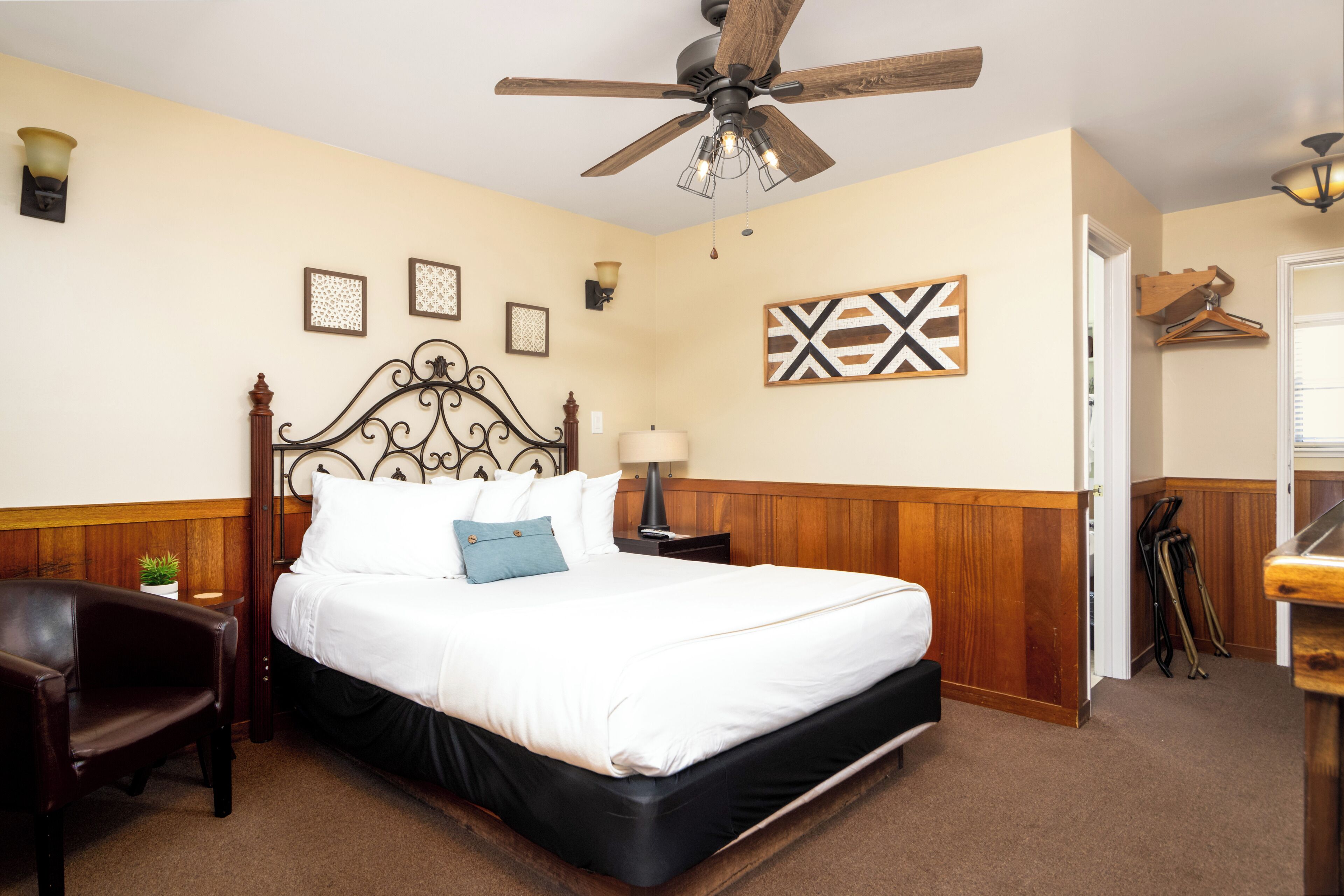 A hotel room with a bed, a ceiling fan, a nightstand, a lamp, and a wooden wall paneling.
