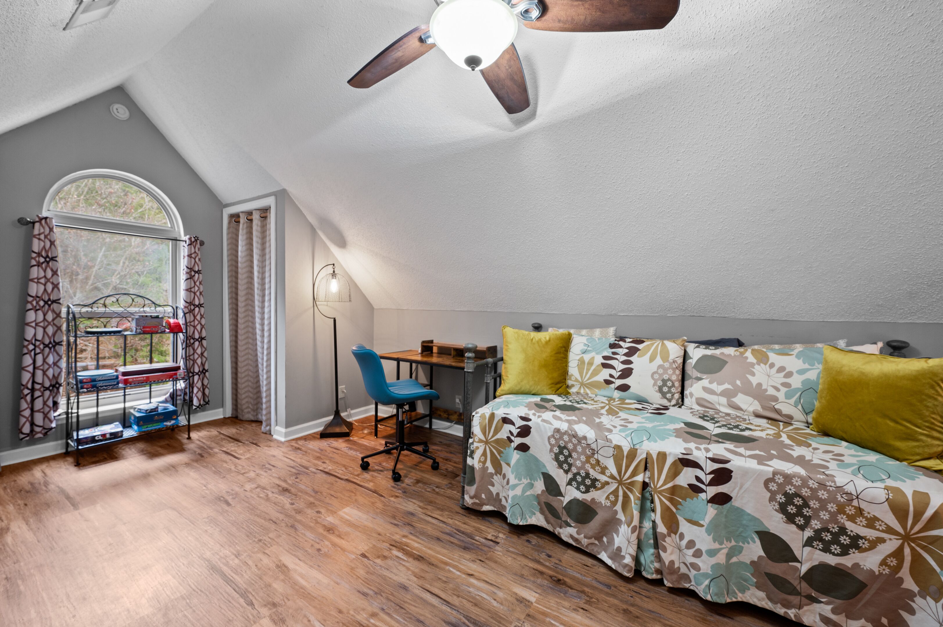 A bedroom with a sloped ceiling, a bed with floral bedding, a desk with a chair, and a window with curtains.