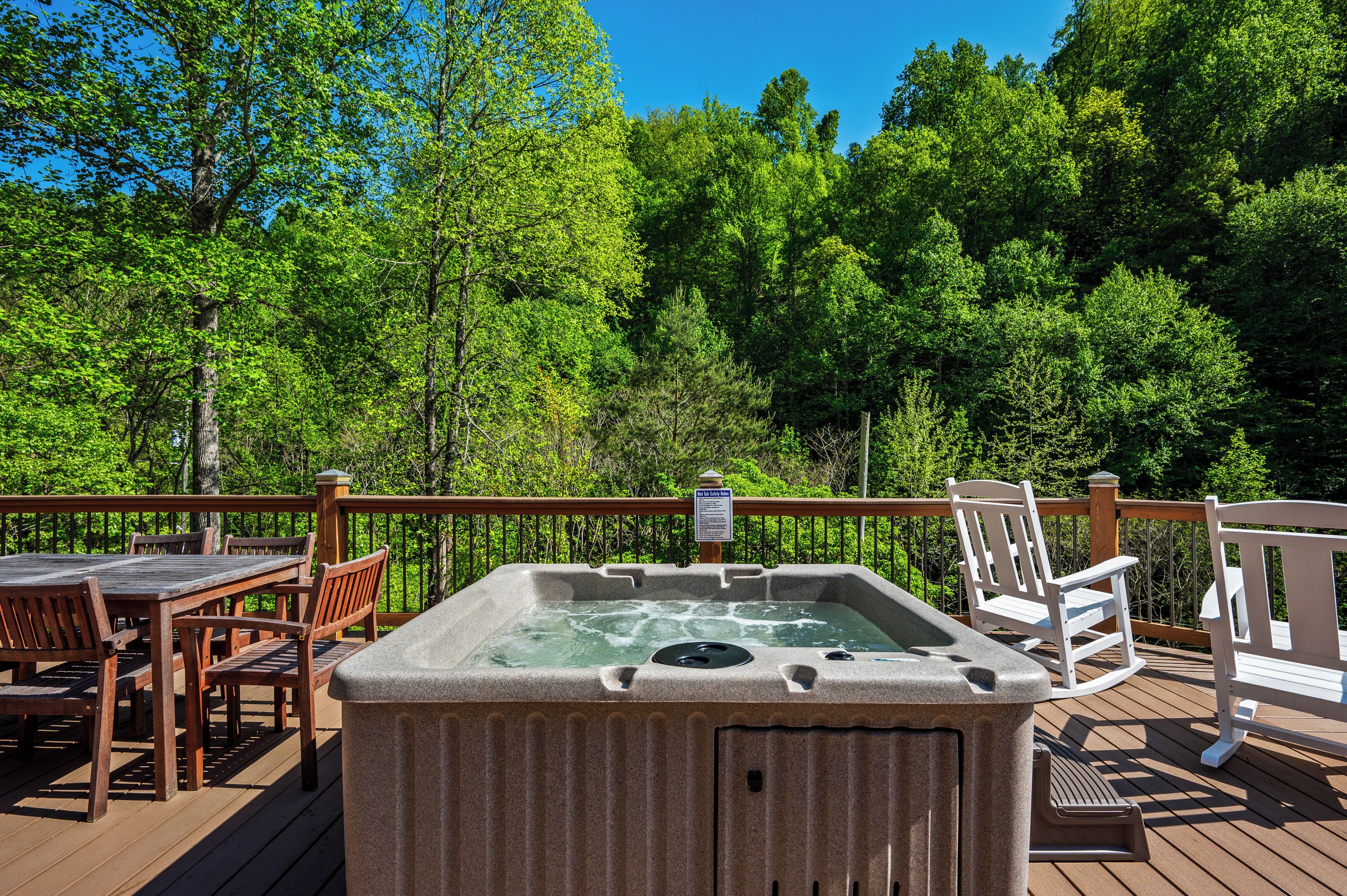 Ein Whirlpool auf einer Holzterrasse mit Holzstühlen und -tischen, umgeben von einer Holzrelung und üppigen Grünpflanzen.