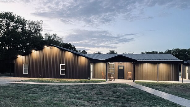 A large, single-story building with a dark exterior and a flat roof. There are two visible doors and windows, and a paved walkway leading to the entrance. - Spacious Lodge on 40 Acres! (Cherryvale)