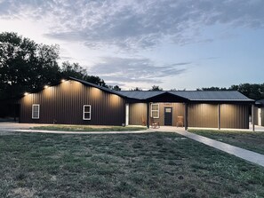 A large, single-story building with a dark exterior and a flat roof. There are two visible doors and windows, and a paved walkway leading to the entrance. - Spacious Lodge on 40 Acres! (Cherryvale)