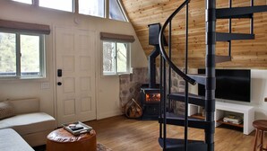 A spacious living room with a wooden ceiling, a black spiral staircase, a stone fireplace, and a large TV on a white stand.