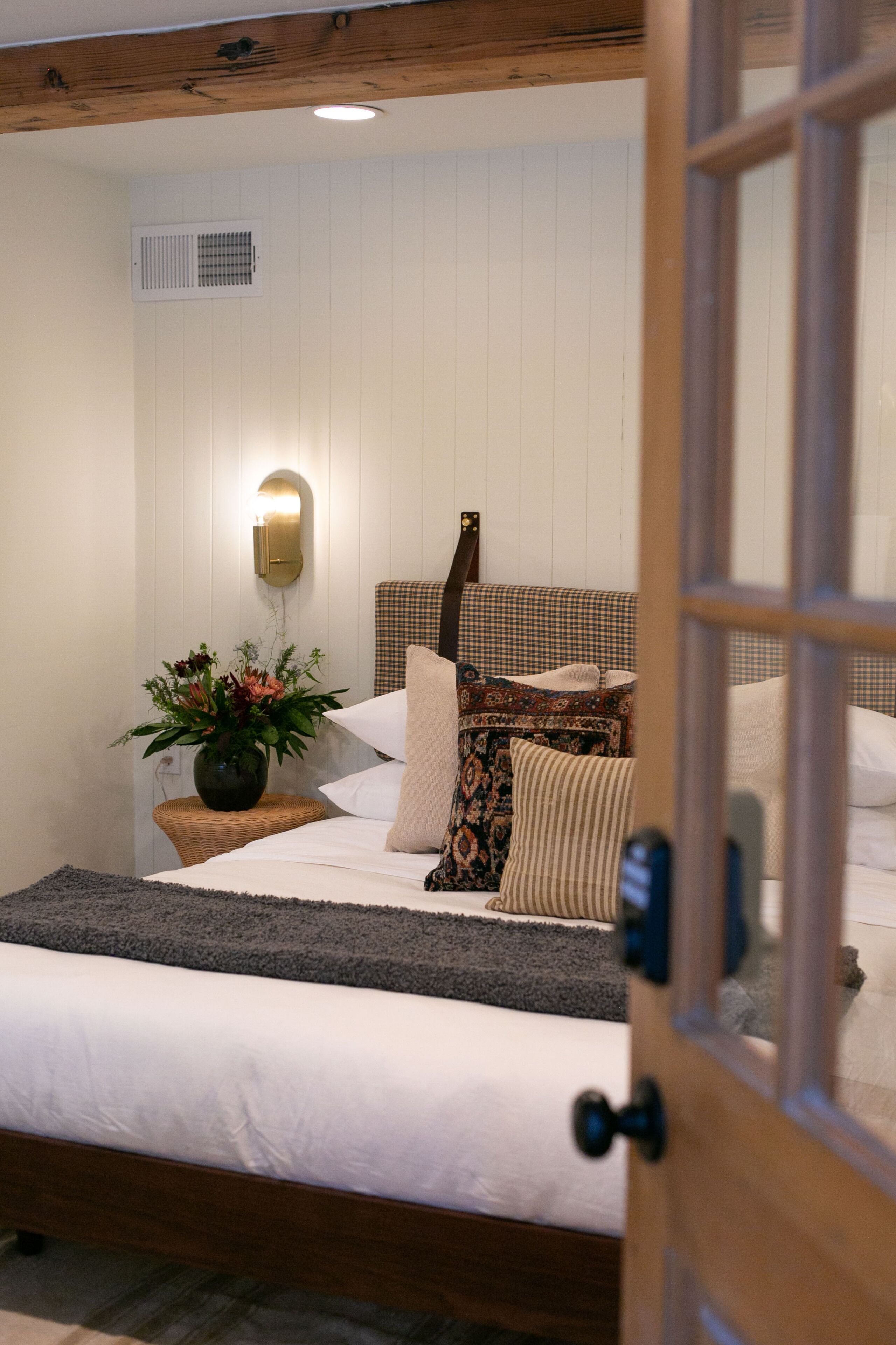 A bedroom with a wooden headboard, a bedside table with a vase of flowers, and a wall-mounted light fixture.