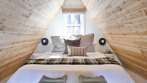 A triangular wooden room with a bed, pillows, a nightstand, and a window.