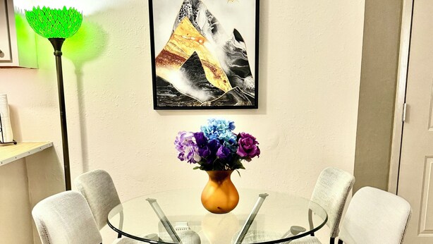 A dining area with a glass table surrounded by four chairs, a vase with flowers, a framed artwork on the wall, and a green floor lamp.