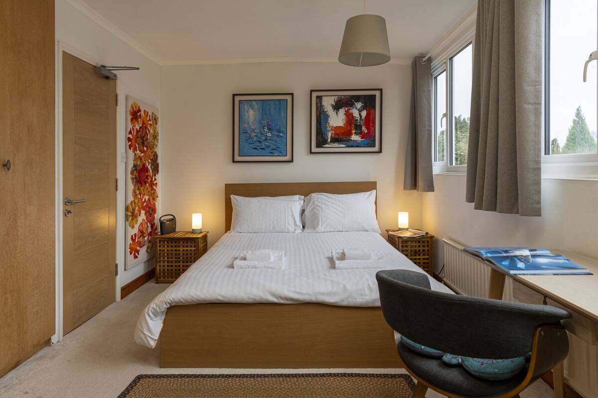 A hotel room with a large bed, two bedside tables, a desk, and a chair. There are two paintings on the wall and a window with curtains.
