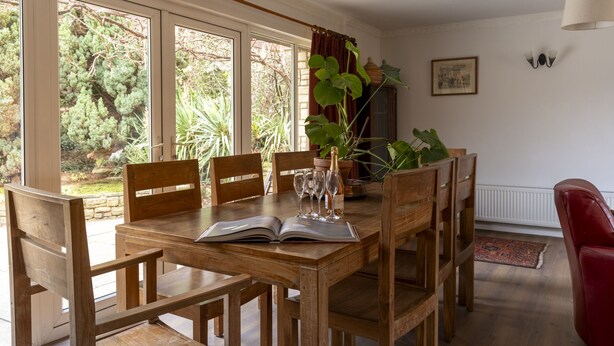 A dining area with a wooden table, chairs, and a view of greenery through large windows. - Sandford Meadow Guest House (Oxford)