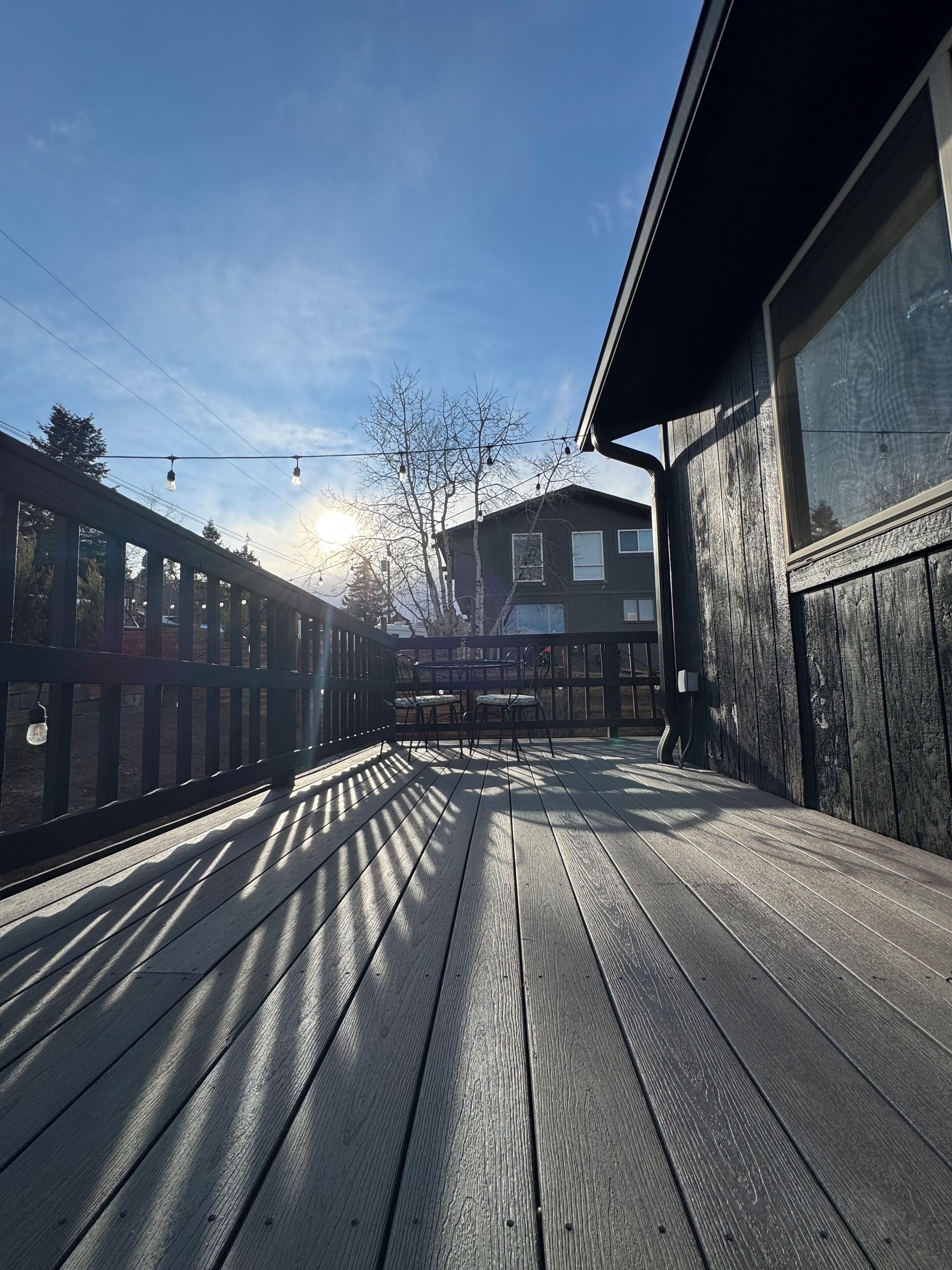 Een houten terras met een tafel en stoelen, een gebouw met een raam, en een heldere lucht.