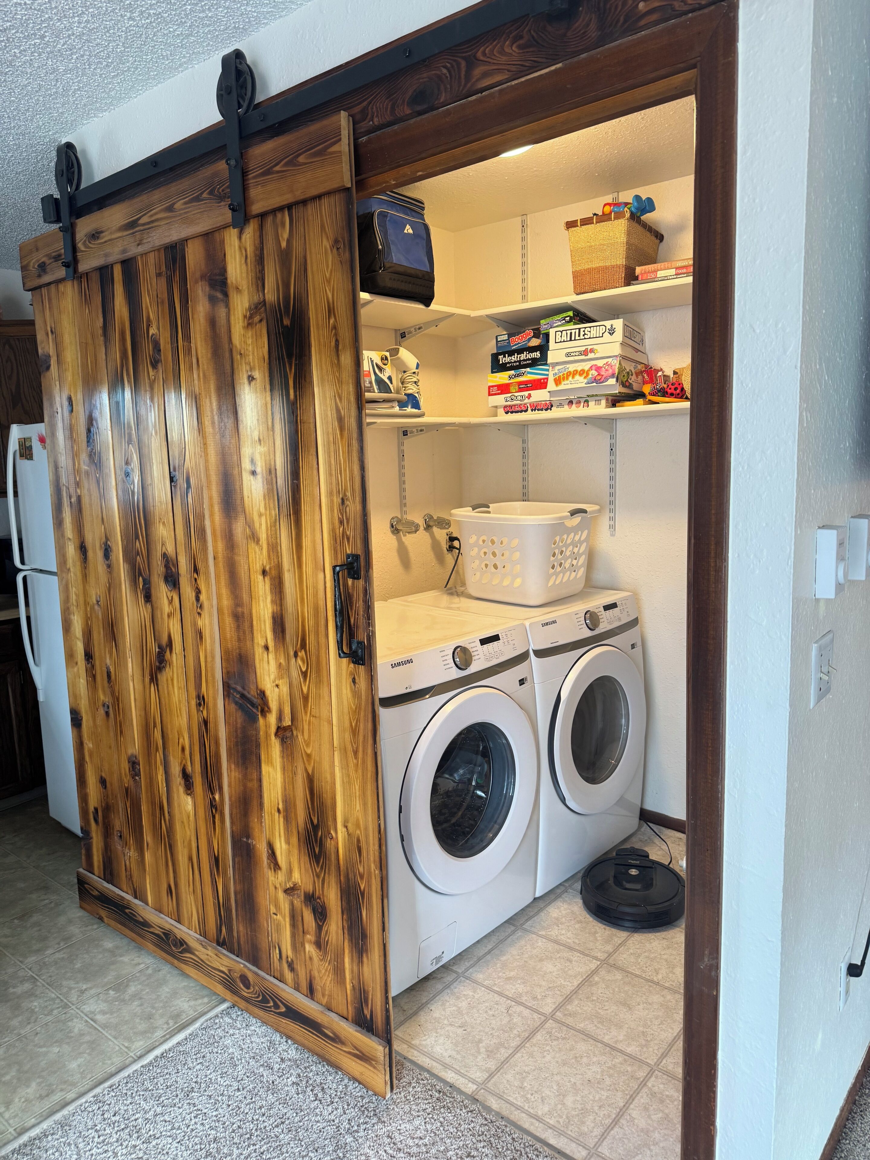 A laundry room with a wooden sliding door, two white front-loading washers, a basket, and shelves with various items.