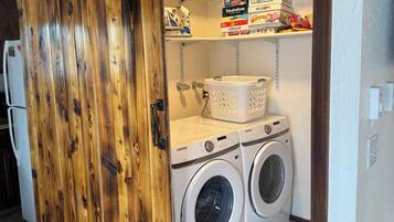 A laundry room with a wooden sliding door, two white front-loading washers, a basket, and shelves with various items.