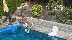 A clear swimming pool with a ladder, two white lounge chairs, and a patio area with a table and chairs.
