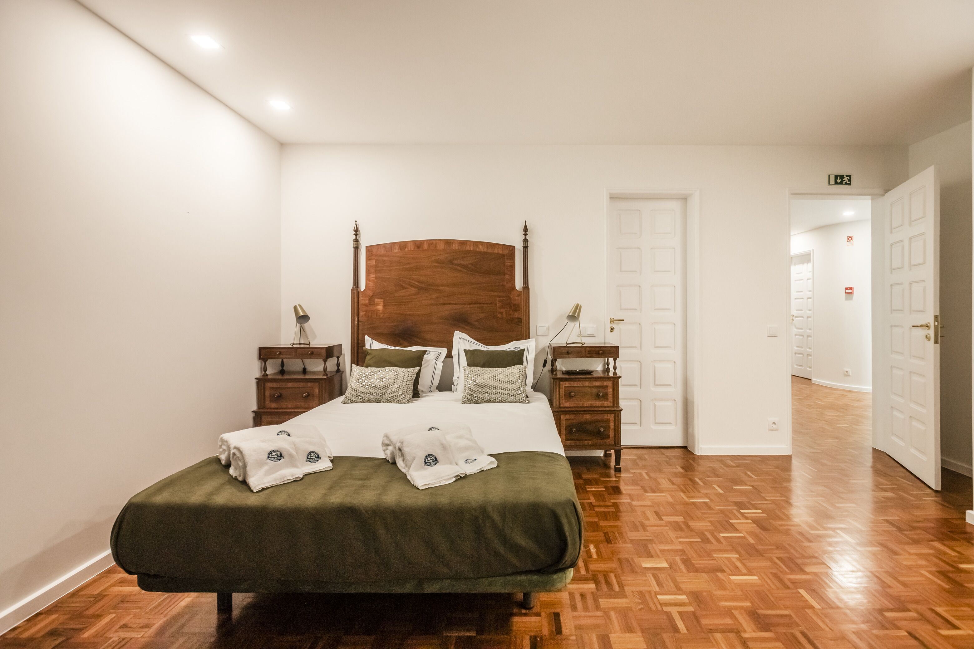 A neatly arranged bedroom with a large bed, wooden headboard, bedside tables, and a door leading to another room.