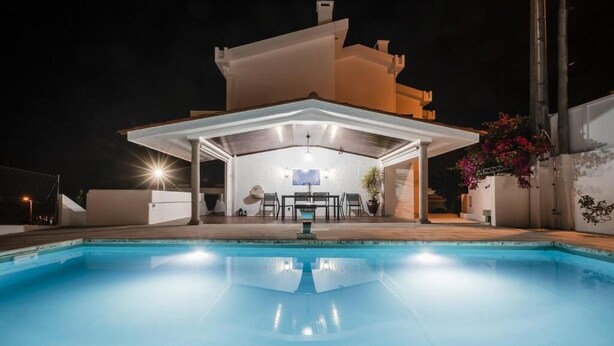 A swimming pool with a covered patio area, a table, and chairs.