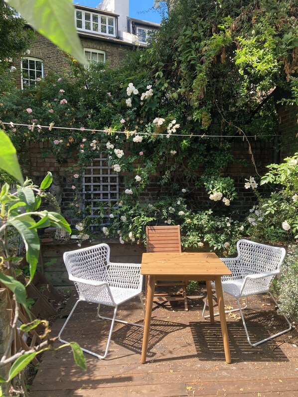 A patio with white wicker furniture, a wooden table, and a brick wall with climbing roses.