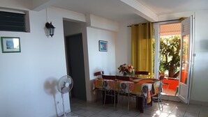 A living room with a sofa, a dining table with chairs, a fan, and a view of the outdoors through a door. - Feet in the water 2 (Les Anses-d'Arlets)