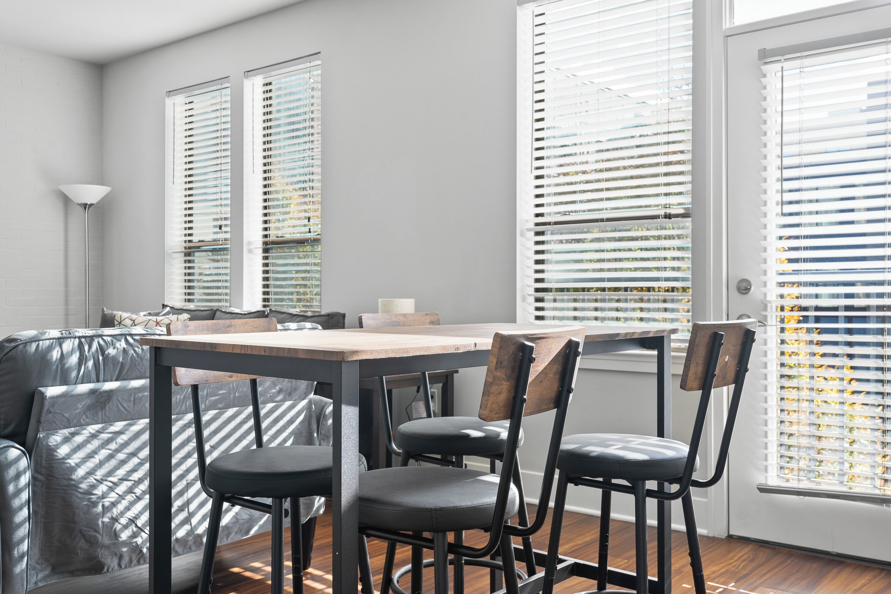 A modern dining area with a wooden table and metal chairs, large windows with blinds, and a sofa with striped cushions.