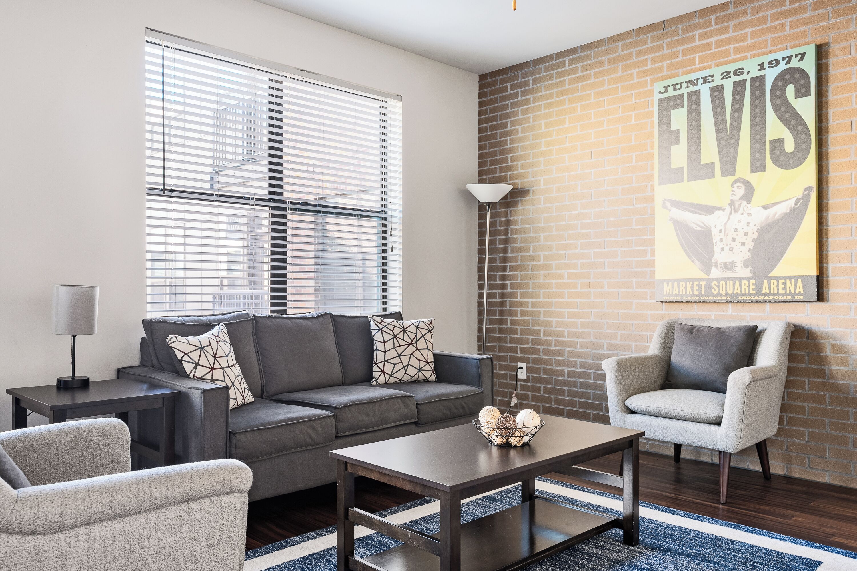 A modern living room with a gray sofa, a coffee table, and a chair. There's a poster on the wall and a lamp on a side table.