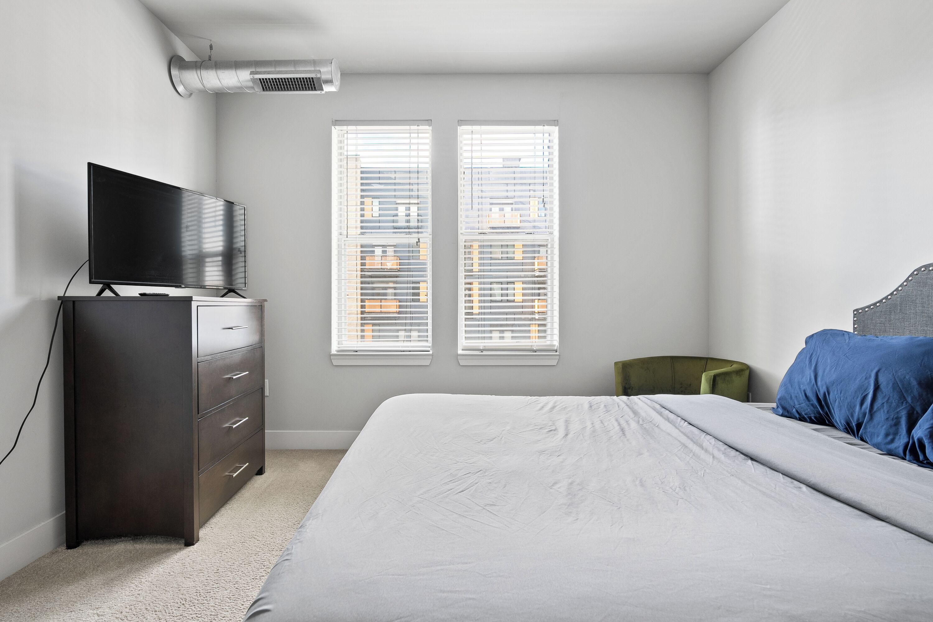 Une chambre à coucher comprenant un lit, une commode, un téléviseur et deux fenêtres équipées de stores.