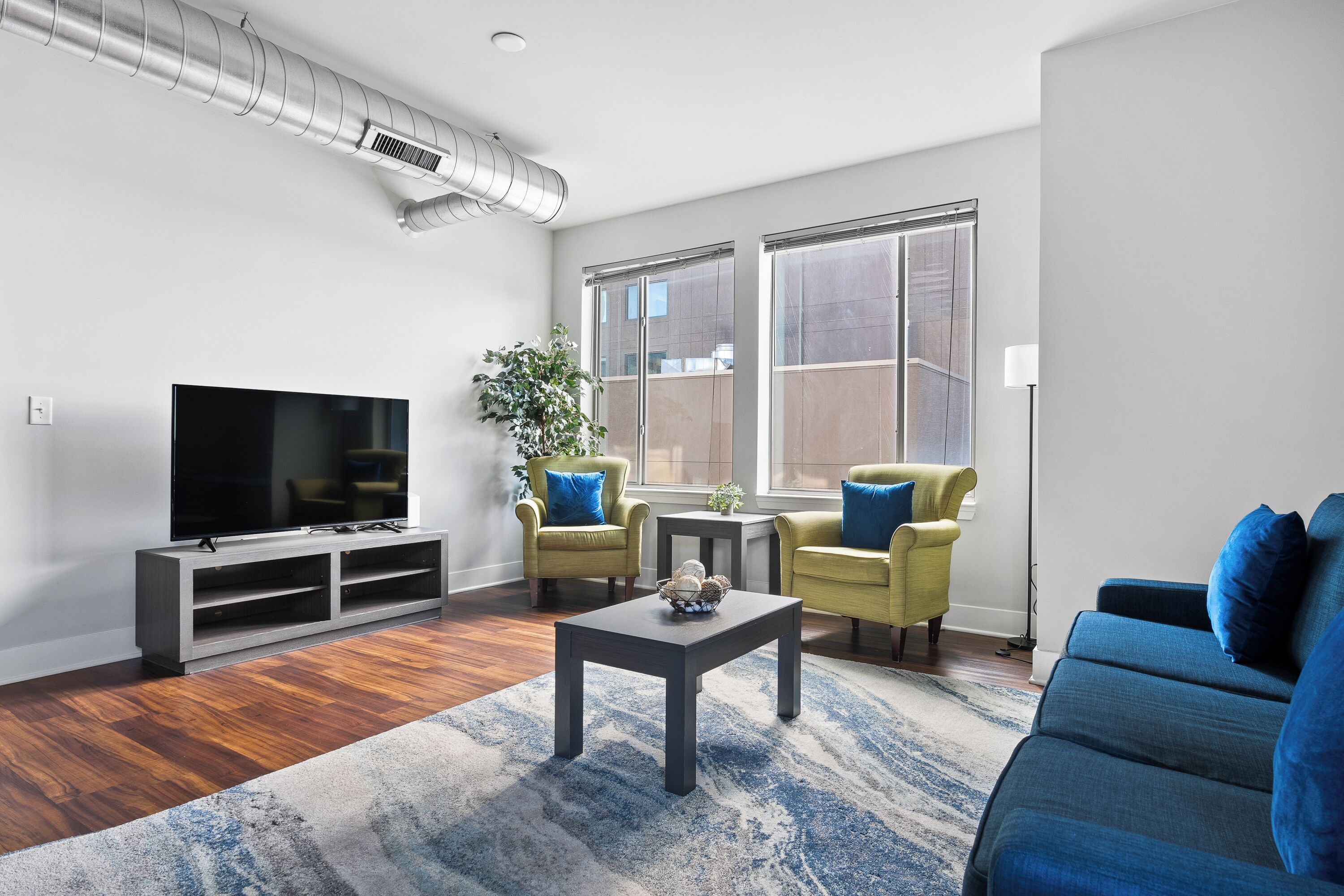 A modern living room with a flat-screen TV, two yellow armchairs, a blue sofa, a coffee table, and large windows.