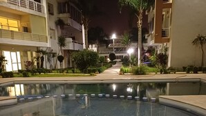 Ein Schwimmbecken mit gefliestem Boden, umgeben von Gebäuden mit Balkonen und Außenbeleuchtung.