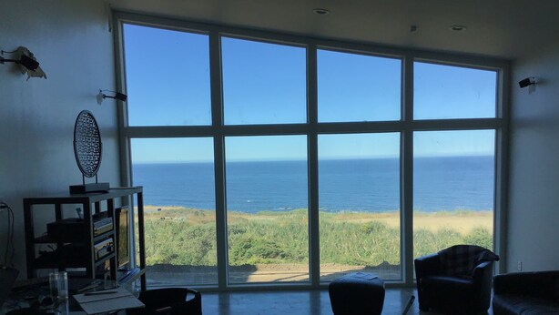 En rummelig stue med stort vindue med udsigt over havet, spiseplads med bord og stole, samt opholdsområde med sofa og skammel.