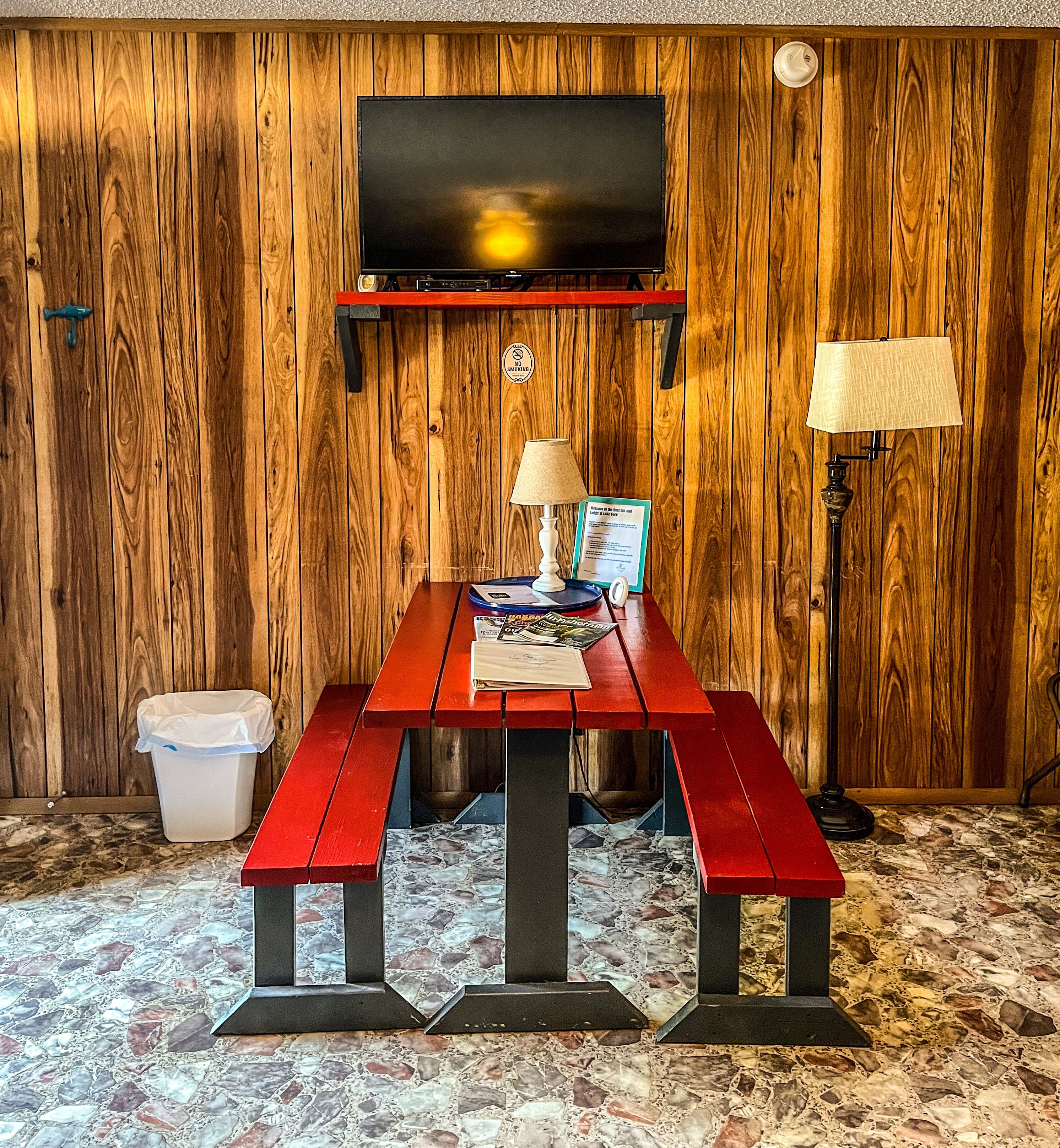 Ein Zimmer mit Holzwandverkleidung, an dem ein Fernseher angebracht ist, eine rote Bank, ein Mülleimer und ein Boden mit Mosaikmuster.