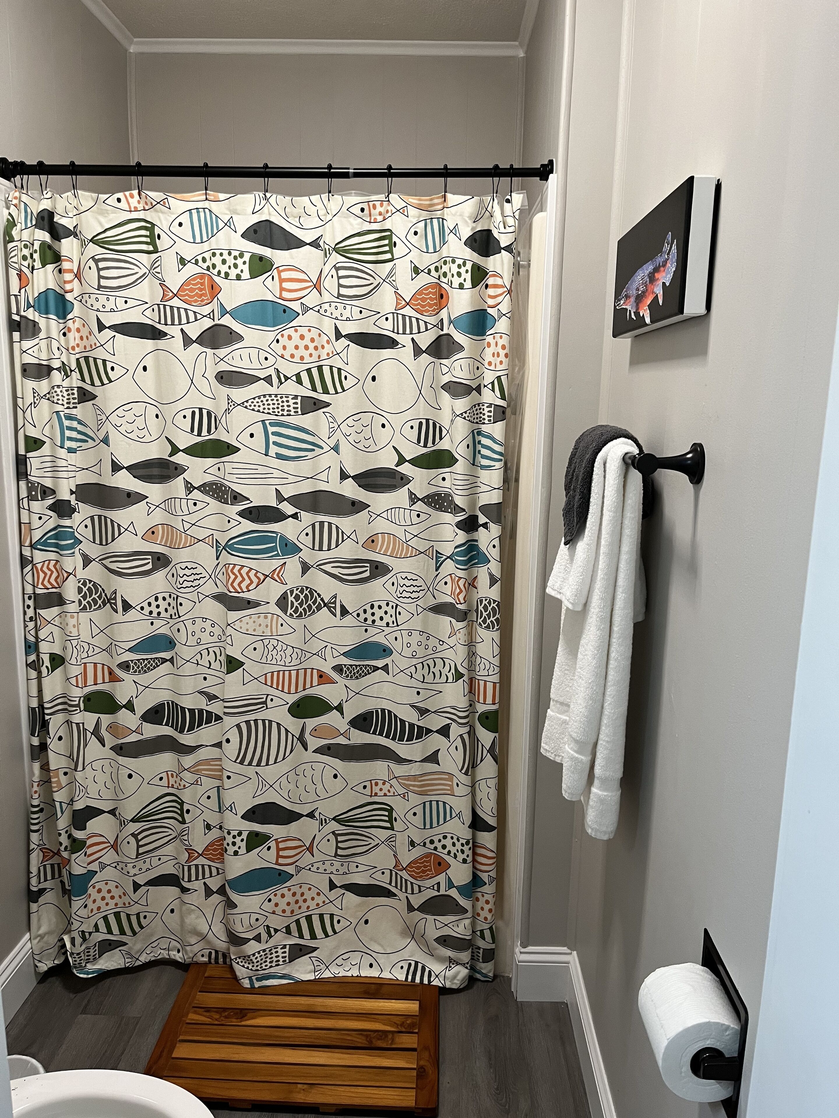 A bathroom with a shower curtain featuring a fish pattern, a wooden bench, a towel rack with towels, a toilet paper holder, and a framed picture on the wall.