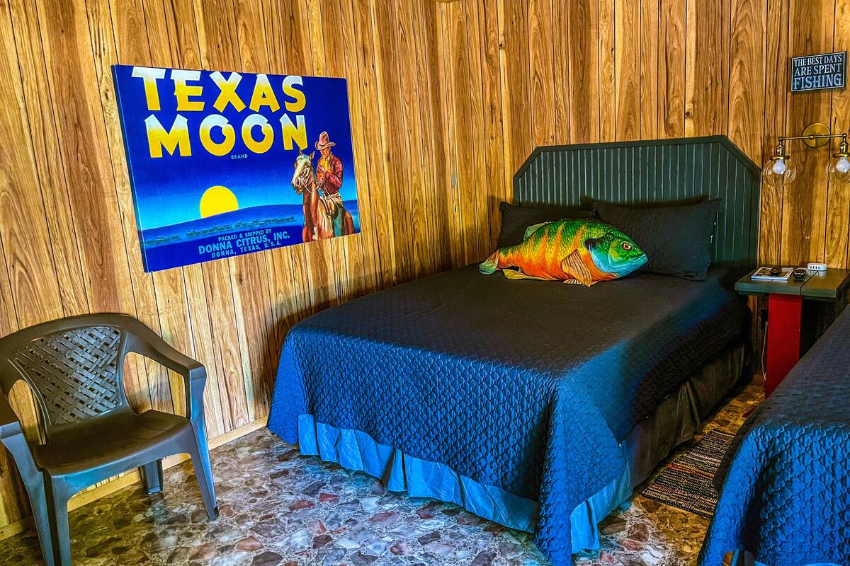 Una habitación con pared de madera, una cama con cubrecama azul, un cartel de Texas Moon, una silla y una pequeña mesa con una lámpara.