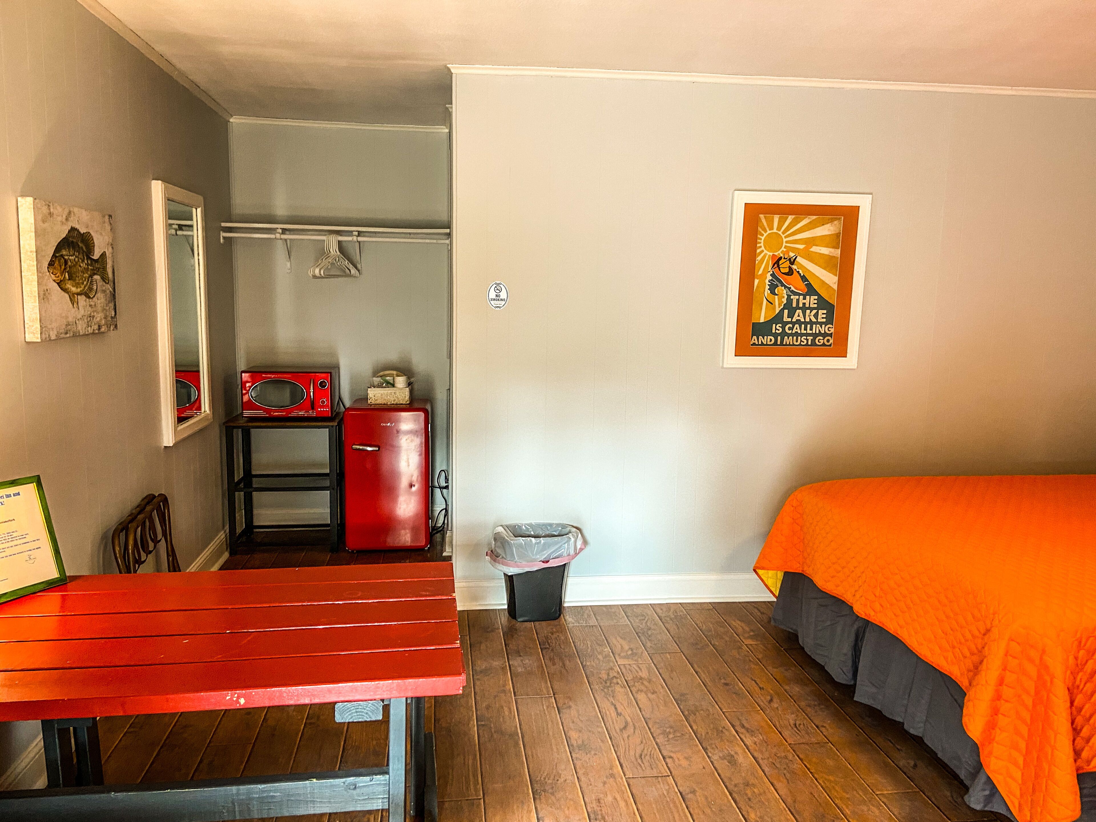 A compact room with a wooden floor, a red table, a bed with an orange blanket, a red refrigerator, a microwave, and a poster on the wall.