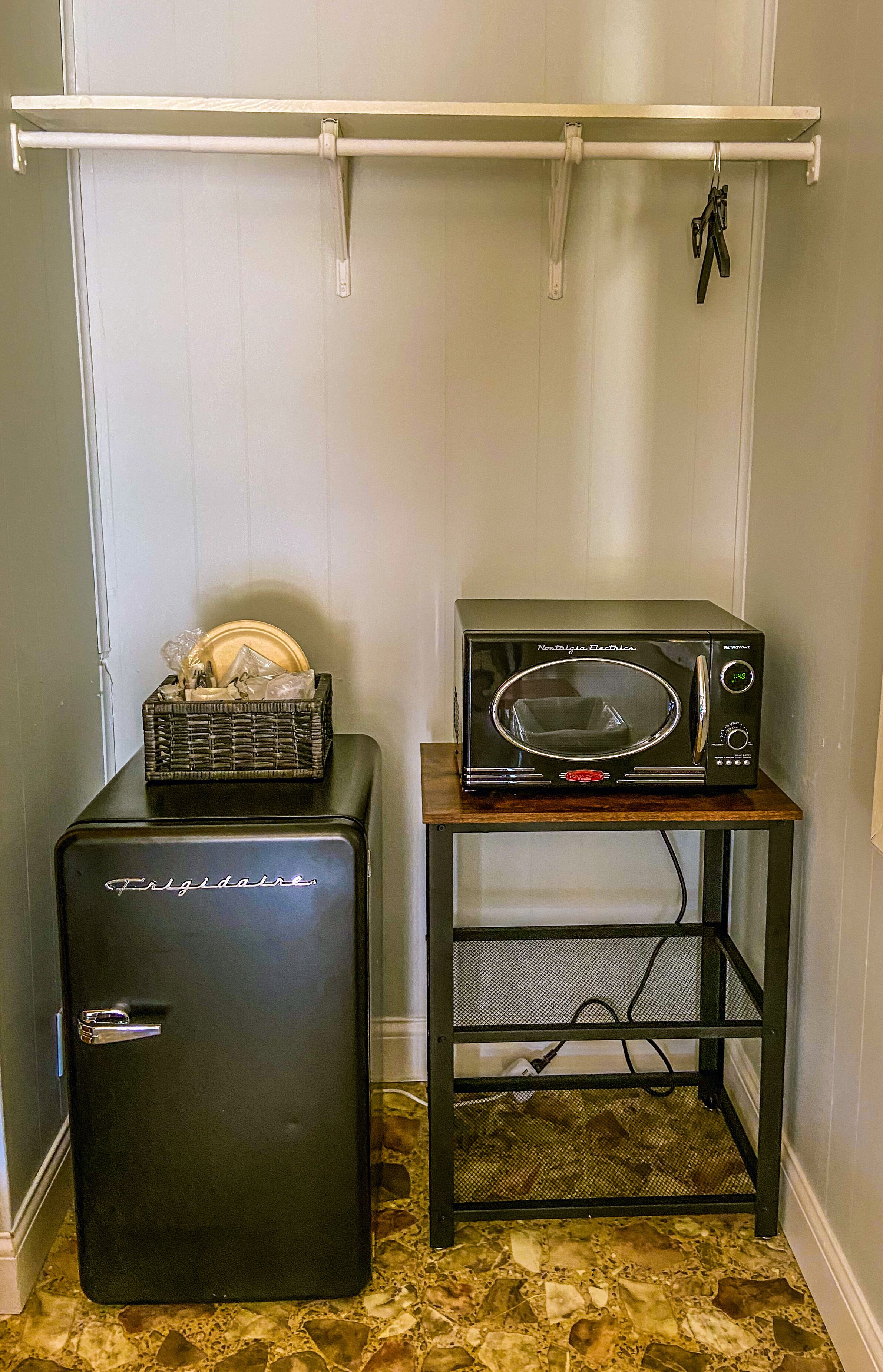 Una kitchenette compacta con un refrigerador negro, un microondas sobre un pequeño estante de madera, y una cesta de platos en el refrigerador.
