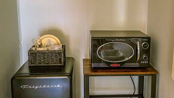 Una kitchenette compacta con un refrigerador negro, un microondas sobre un pequeño estante de madera, y una cesta de platos en el refrigerador.