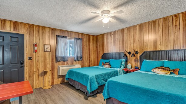 A room with two beds, wooden walls, a ceiling fan, a fire extinguisher, and a window with curtains.