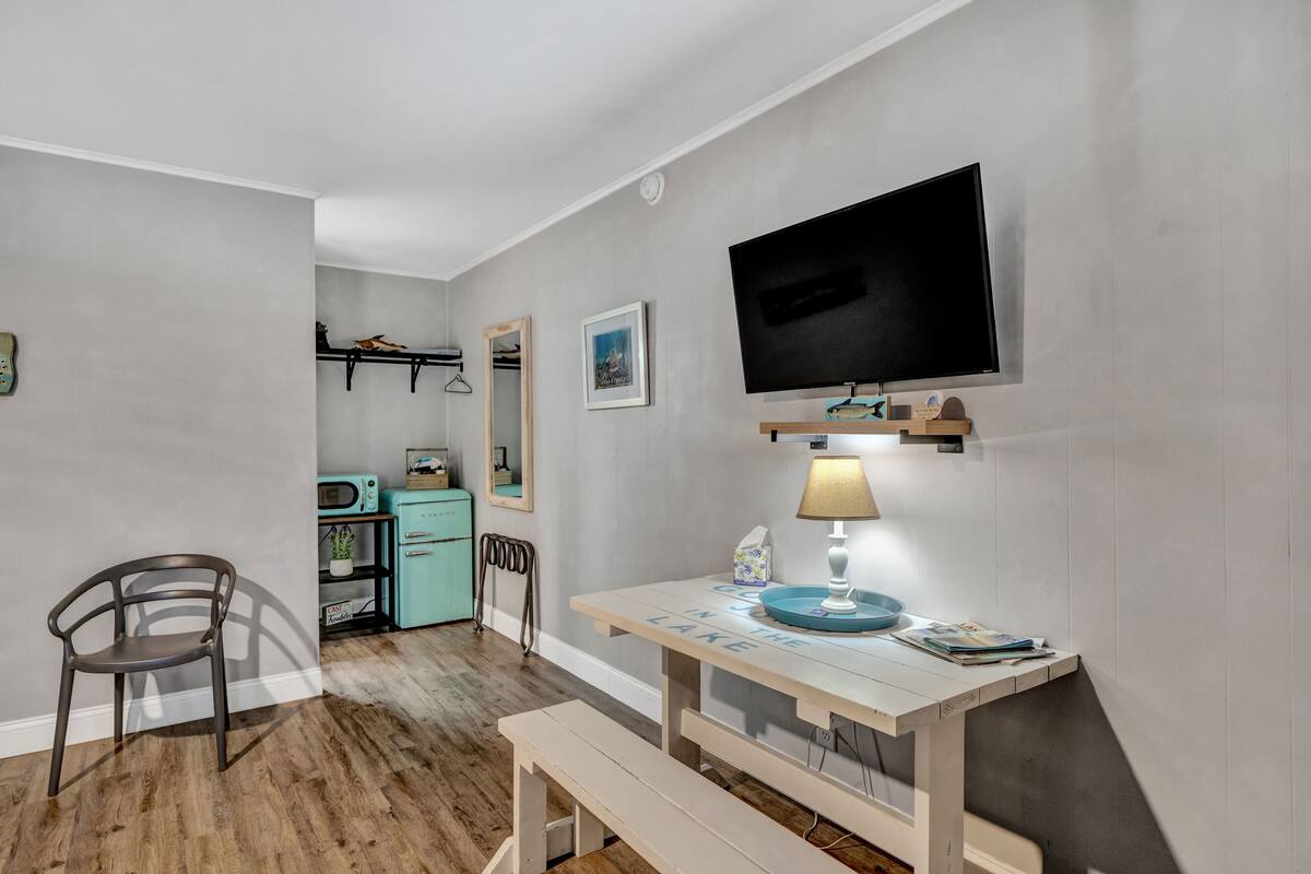 Una habitación compacta con suelo de madera, una mesa pequeña con lámpara, televisión montada en la pared y una silla.