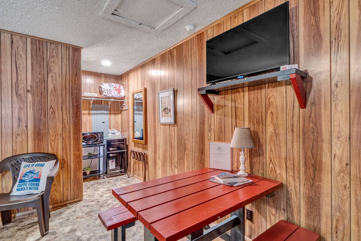 Habitación compacta con paneles de madera, un televisor montado en un estante de madera, una mesita con lámpara y una silla con cojín.