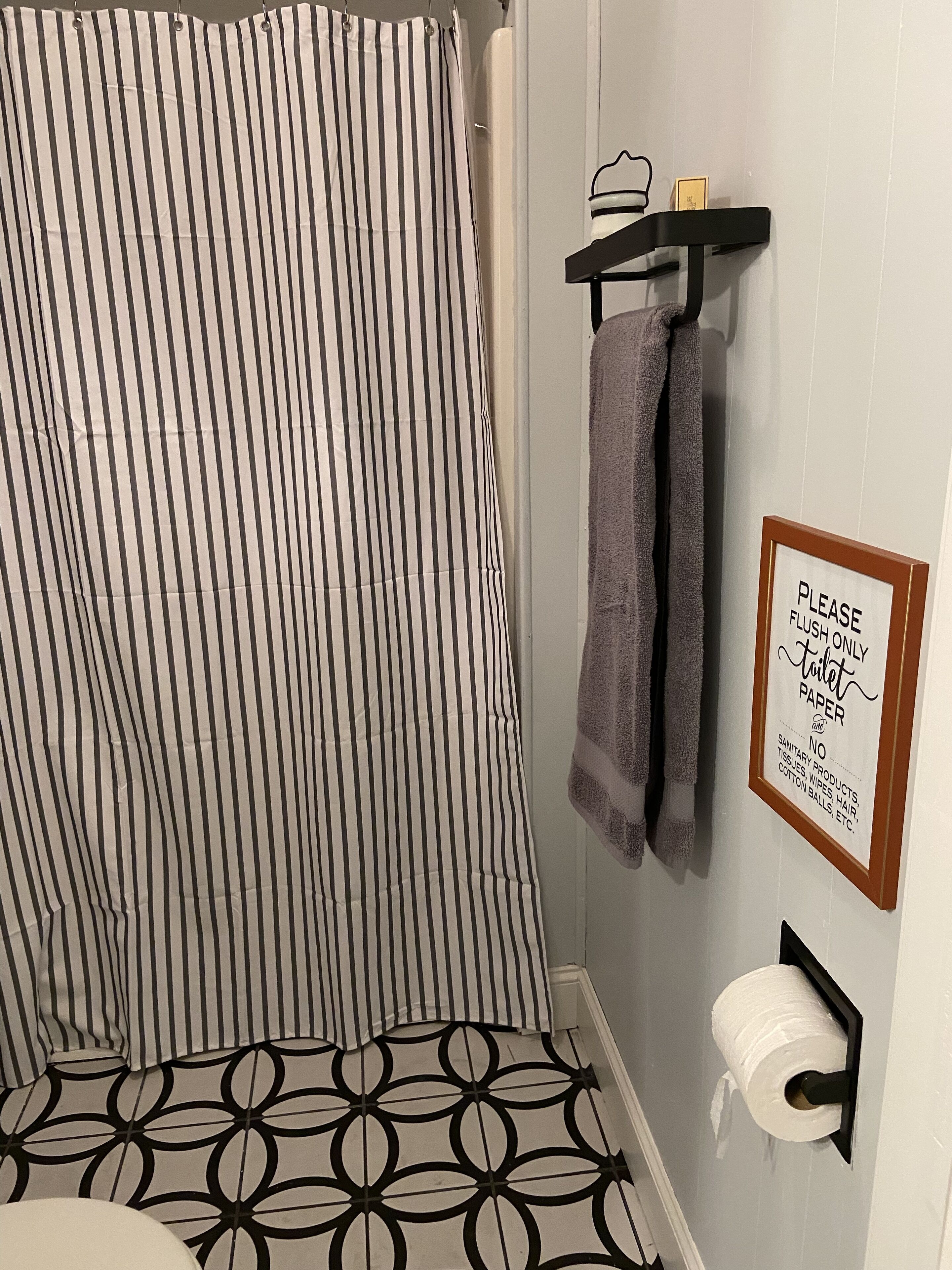 A bathroom with a striped shower curtain, a towel rack with towels, a sign prohibiting sanitary products, and a toilet paper holder.