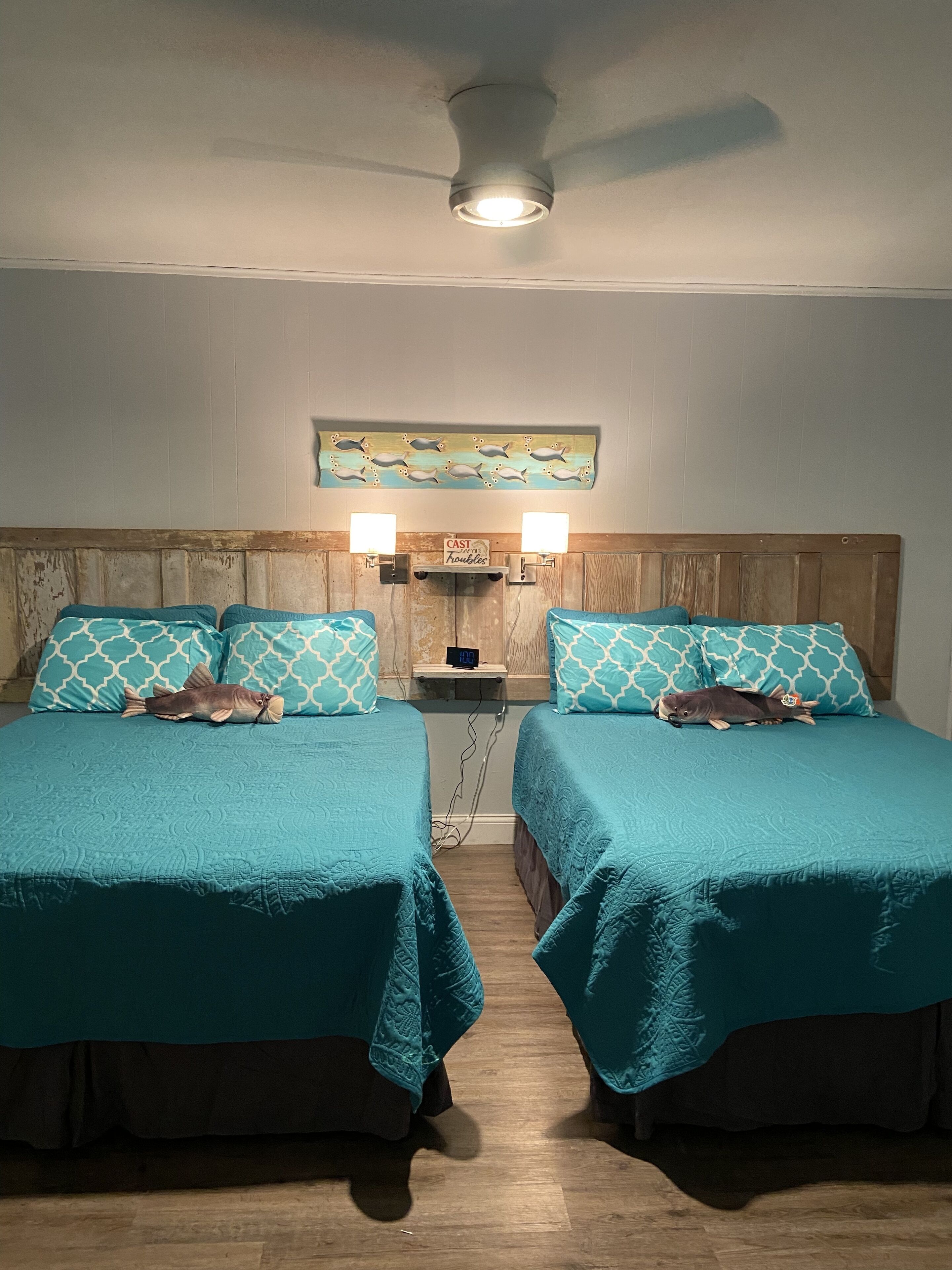 A room with two beds, each with teal bedding and patterned pillows, a wooden headboard, a wall-mounted shelf with a clock, and a ceiling fan.