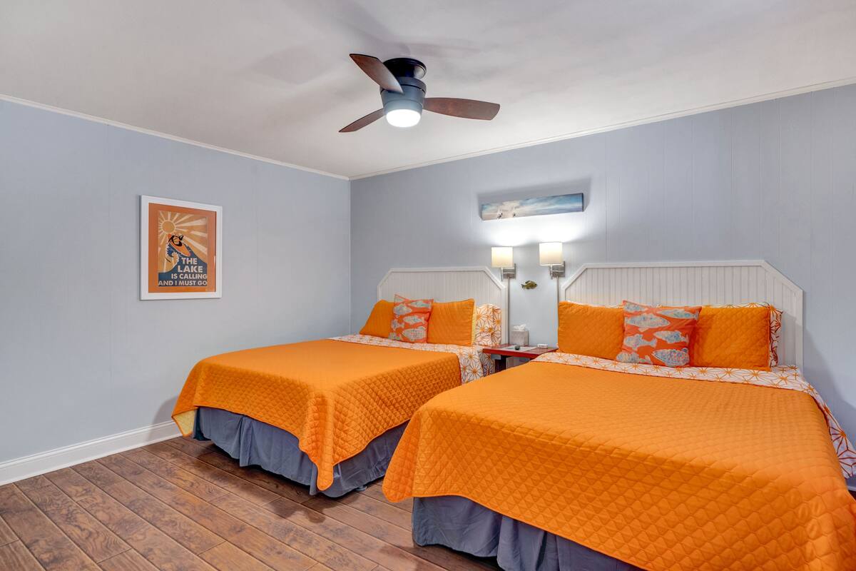 Una habitación con dos camas, cada una con una colcha naranja y almohadas a juego. Hay un ventilador de techo, una lámpara fijada a la pared y un cuadro enmarcado.