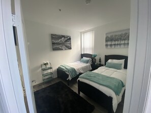 Two single beds with dark headboards and white bedding, a large window, and a decorative shelf with a vase of flowers.