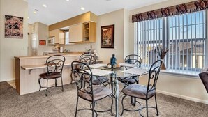 A dining area with a round table and six chairs, a kitchen area with cabinets and appliances, and a large window with blinds. - Lux 2BR Condo w/Jacuzzi, fast Wifi, Clubhouse, Gym (Branson)