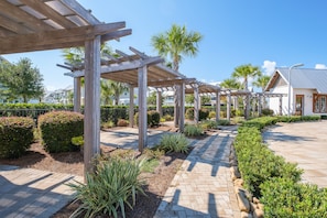 A covered outdoor area with wooden pergolas, a paved walkway, and landscaped greenery including palm trees and shrubs.