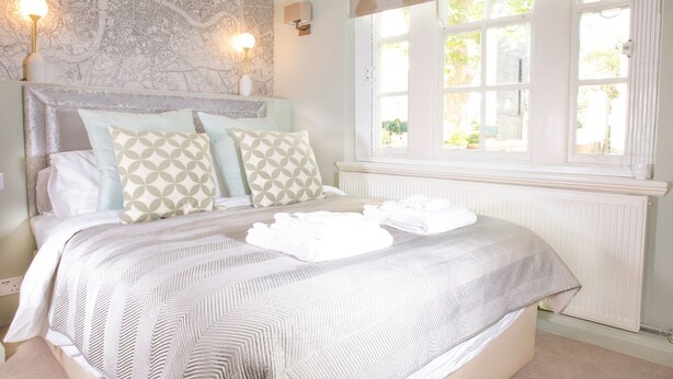 A neatly made bed with patterned bedding, a headboard with decorative molding, and a large window with a view of greenery. - Super Stylish Living in the Best Location in the Heart of Kensington (London)