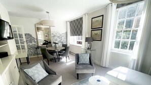 A modern living room with a sofa, armchairs, a dining table, and a coffee table. There is a large window with curtains, a fireplace, and framed pictures on the wall. - Super Stylish Living in the Best Location in the Heart of Kensington (London)