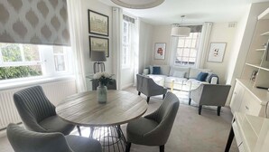 A modern living room with a round wooden table, grey upholstered chairs, a grey sofa, and a white armchair. There are framed pictures on the walls, a vase with white flowers on the table, and a window with white curtains. - Super Stylish Living in the Best Location in the Heart of Kensington (London)