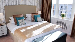 Un letto rifatto con cura, dotato di una testiera, diversi cuscini, un comodino con una lampada e una finestra che offre vista sugli edifici.