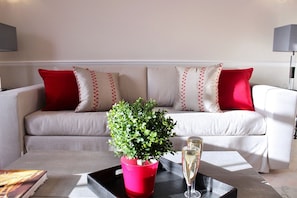 A living room with a sofa, red and white pillows, a potted plant, and two glasses of champagne on a tray.