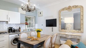 A modern kitchen with white cabinets, a coffee maker, and a washing machine. There is a dining table with chairs and a vase of yellow flowers. A large ornate mirror hangs on the wall.