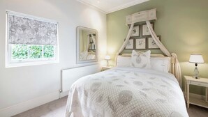 A bedroom with a large bed, a bedside table with a lamp, a wall-mounted mirror, and framed botanical prints.