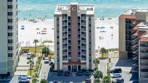 Un hotel con una torre centrale, una spiaggia sullo sfondo e un'area parcheggio con auto.