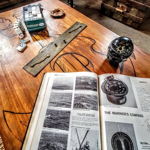Une table en bois sur laquelle repose un livre ouvert à une page intitulée « La boussole du marin », illustrant les forces du vent et présentant une boussole.
