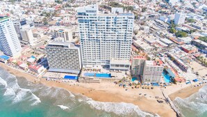 A coastal cityscape with a prominent hotel featuring multiple floors, a beach with umbrellas, and a clear view of the ocean.